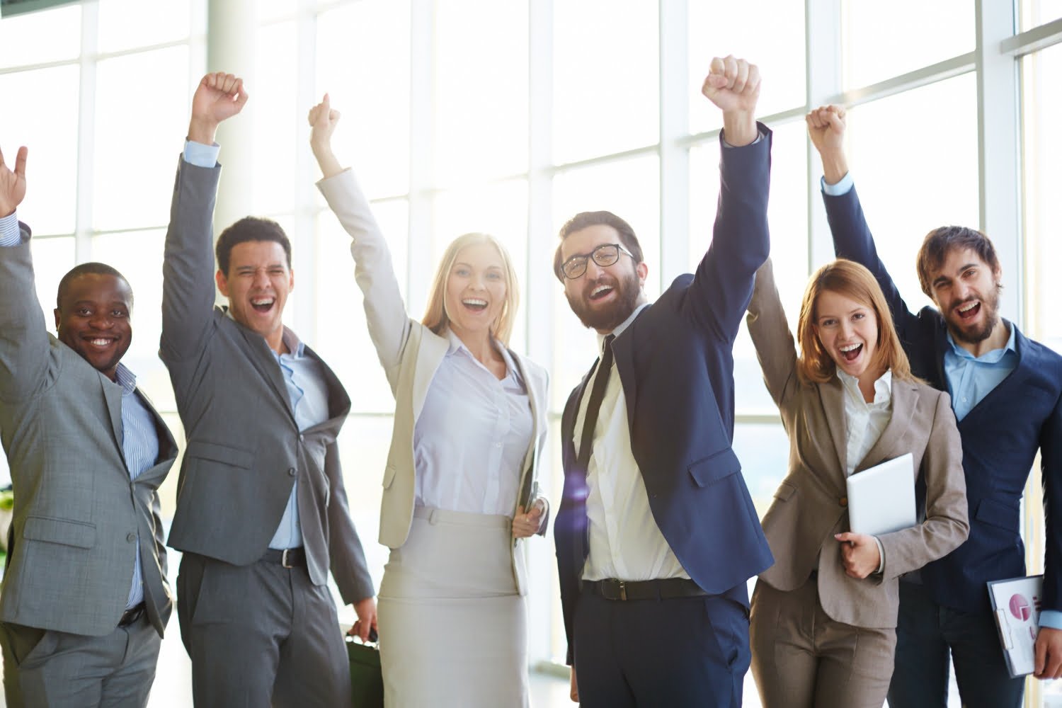 A group of six diverse professionals in business attire, smiling and raising their fists in celebration, stand in a bright office space. People are happy for getting employment visa