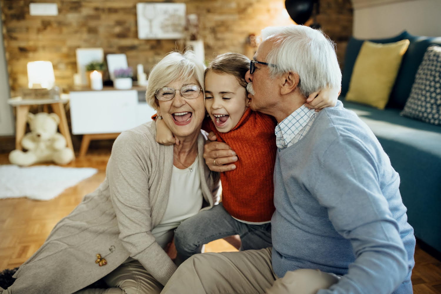 Elderly couple and young girl share a joyful moment on the floor, with the grandfather kissing the girl’s cheek. Warm, cozy living room setting. Happy family getting parent visa