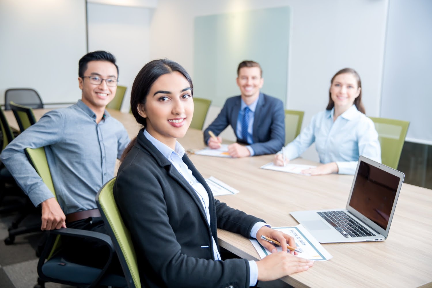 Four skilled professionals in a modern conference room smile towards the camera. A woman in a blazer sits at the table's head with a laptop, exuding confidence.