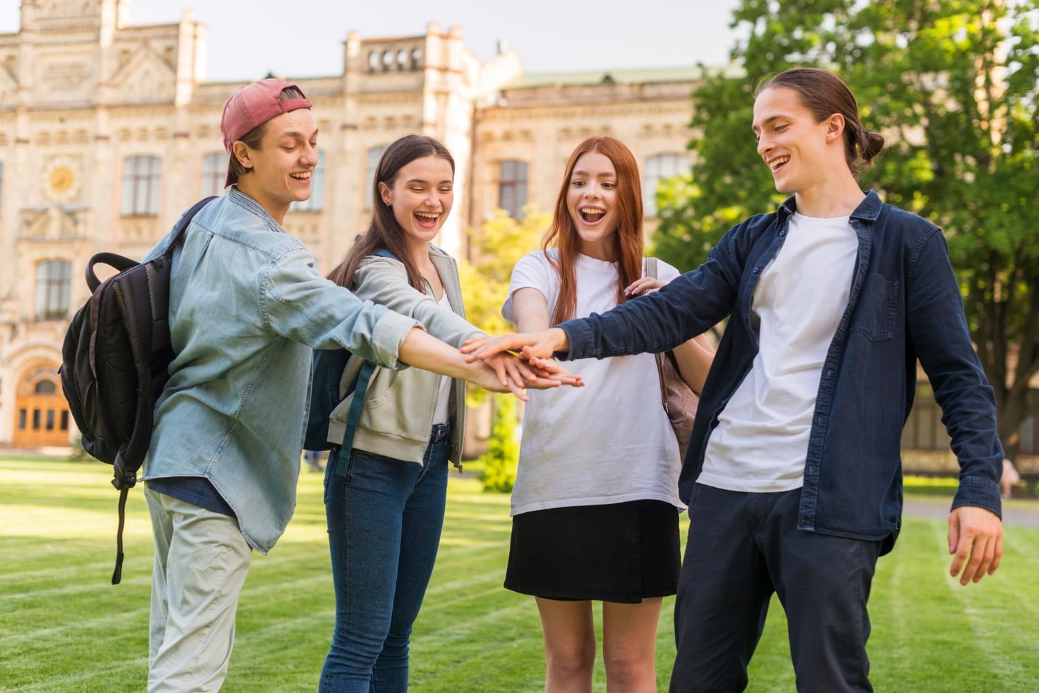 Four students stand in a circle on a lawn, hands stacked cheerfully in the center. They're laughing, with a historic building and trees in the background. They are here through student visa