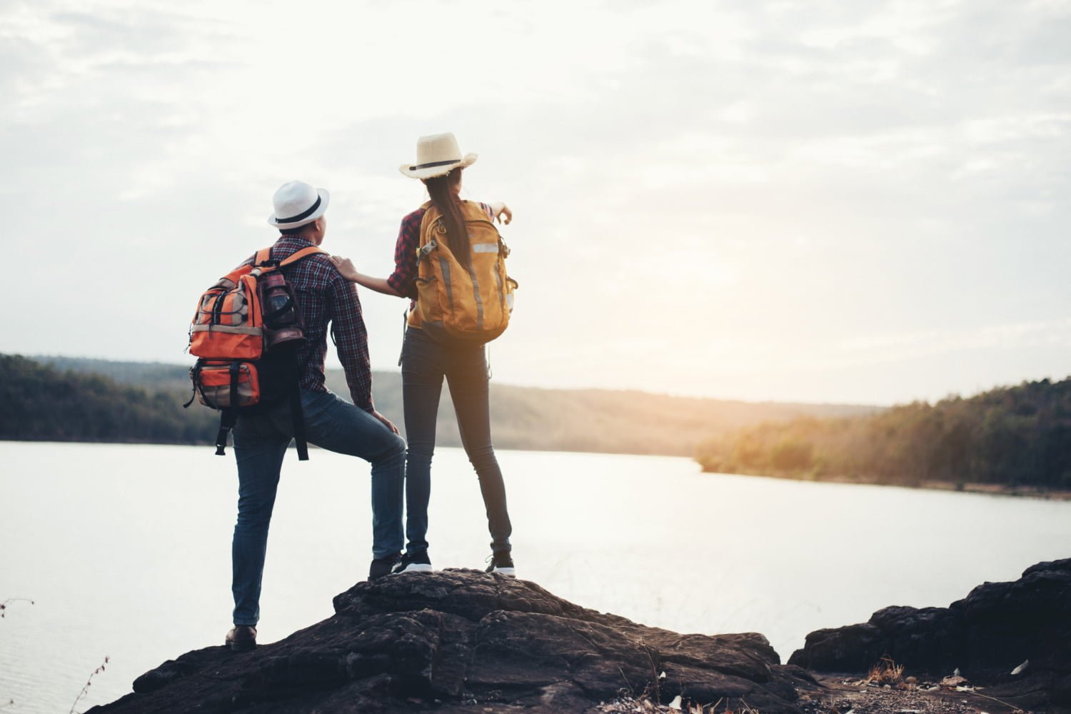 Two tourist with backpacks and hats stand on a rocky cliff, gazing at a serene lake under a cloudy sky, conveying adventure and tranquility. Happy travelling tourist visa